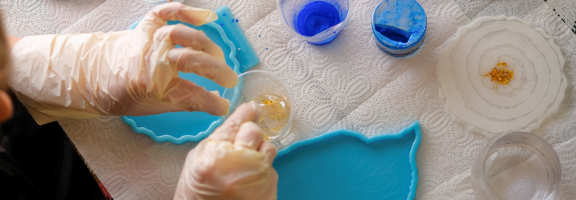 Gloved hands using resin to create a coaster with craft supplies surrounding a cup of resin.