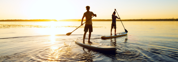 Two boys dressed in shirts and shorts standing up on separate paddle boards on a water as the sun rises behind them.